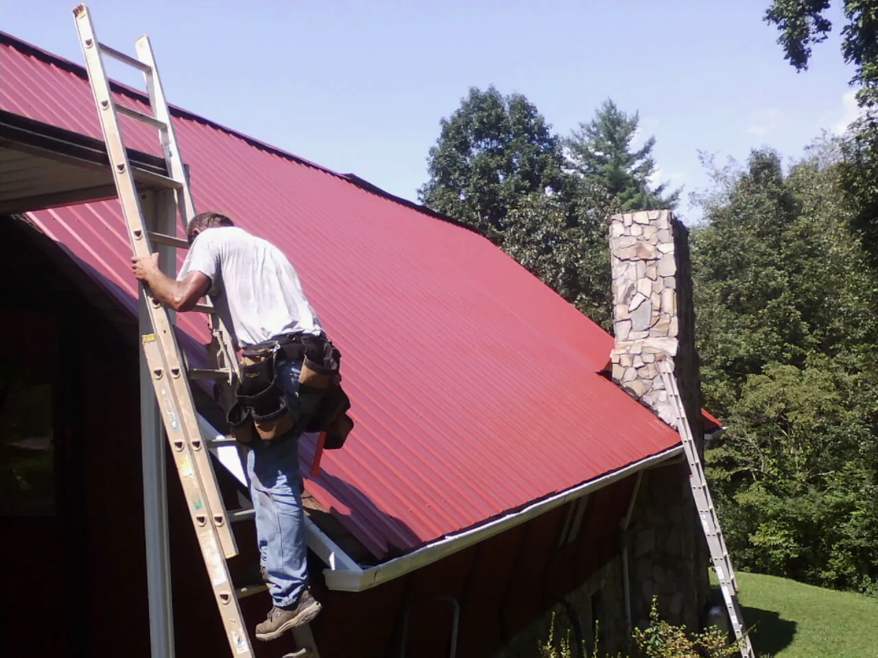Roofer on a ladder beside a red metal roof and stone chimney — Soffit & Fascia Repair by Roofing Brothers in Cavour